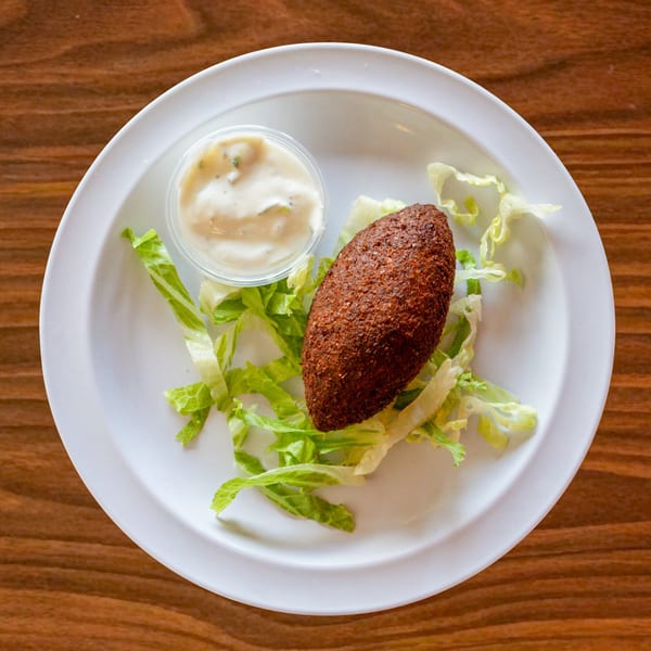A single kibbeh, a container of sauce, and lettuce on a white plate from Zest Mediterranean Cuisine.