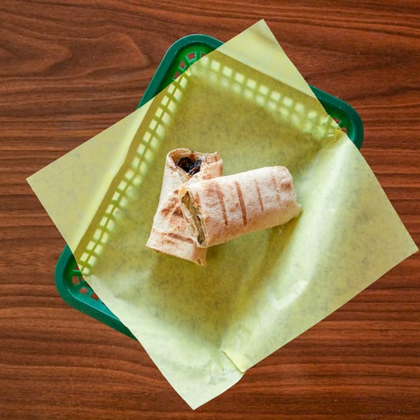 Two Mediterranean wraps with grilled marks, one open, in a green basket with yellow paper on a wooden table.